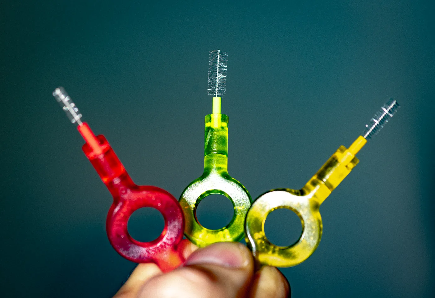 Trois brossettes interdentaires de tailles différentes, rouge, verte et jaune, tenues à la main